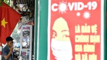 A propaganda poster about preventing the spread of coronavirus is seen on a wall while a man smokes a cigarette on a Hanoi street.